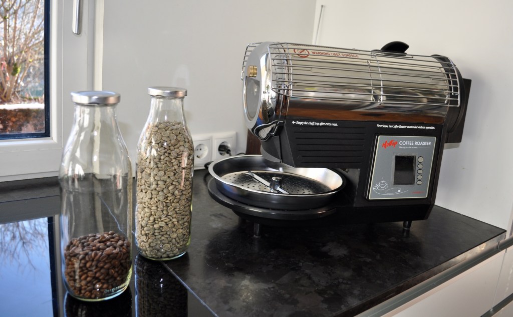 Image showing a home coffee roaster manufactured by the US company HotTop, along with sampes of roasted and unroasted coffee beans in glass bottles placed next to it.