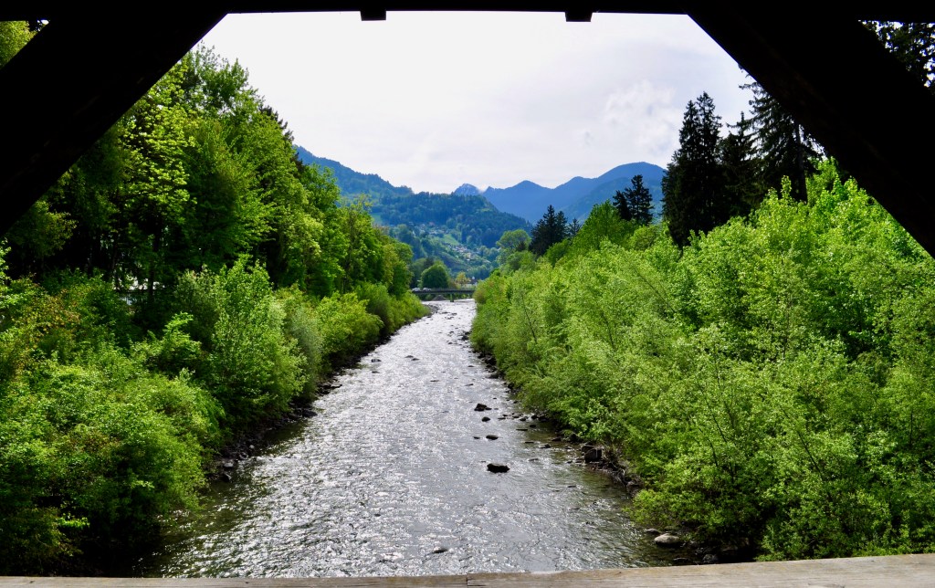 This is a photograph looking upstream of a small mountain river from a covered wooden bridge across it. It was taken in May, so the banks are verdant green with new growth. On the horizon you can see mountains, suggestive of the steep-sided V-shaped valley that this stream drains.