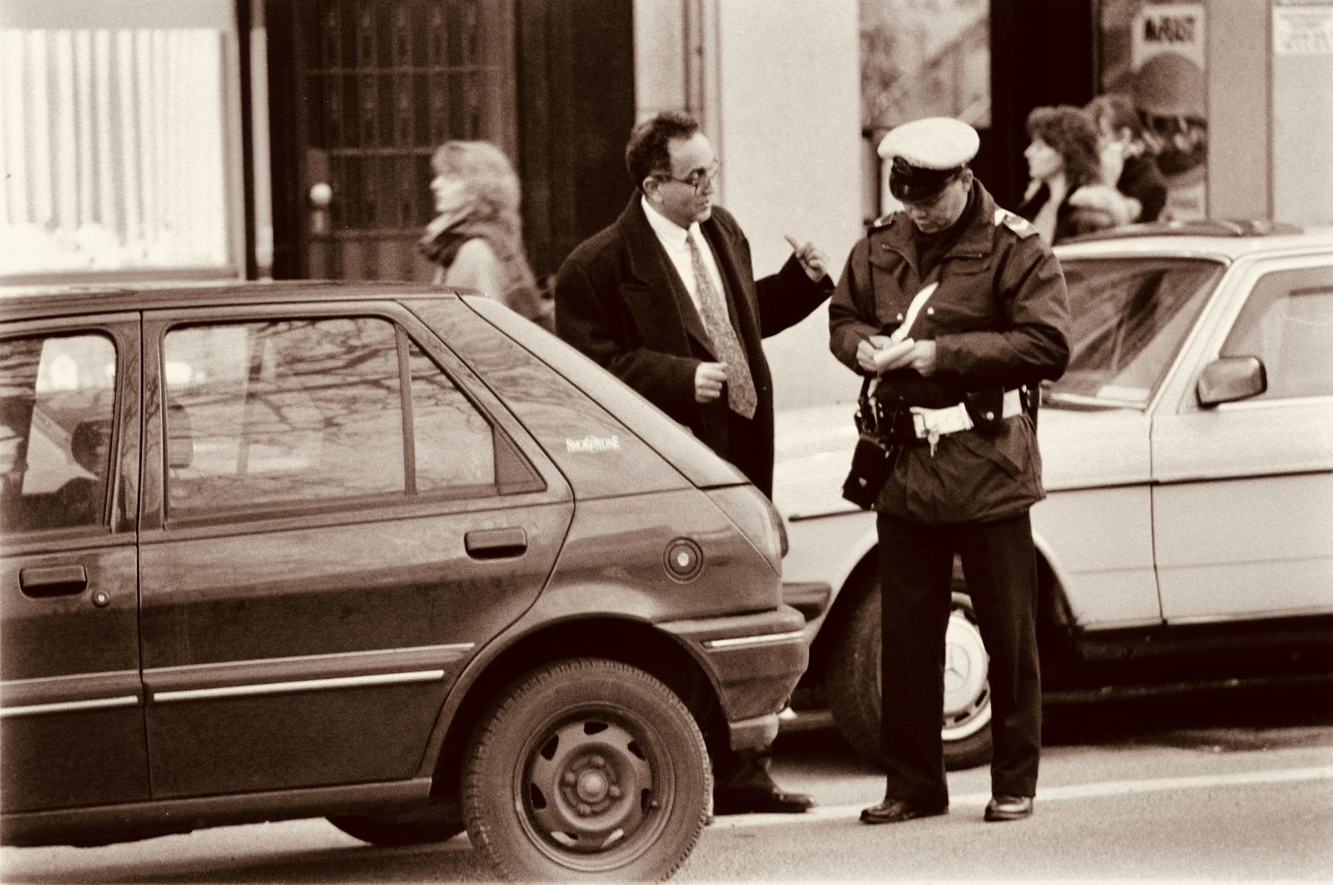 A monochrome photograph showing a parking attendant standing behind a presumably illegally parked car, writing out a ticket for the owner, who is standing next to him talking, explaining.